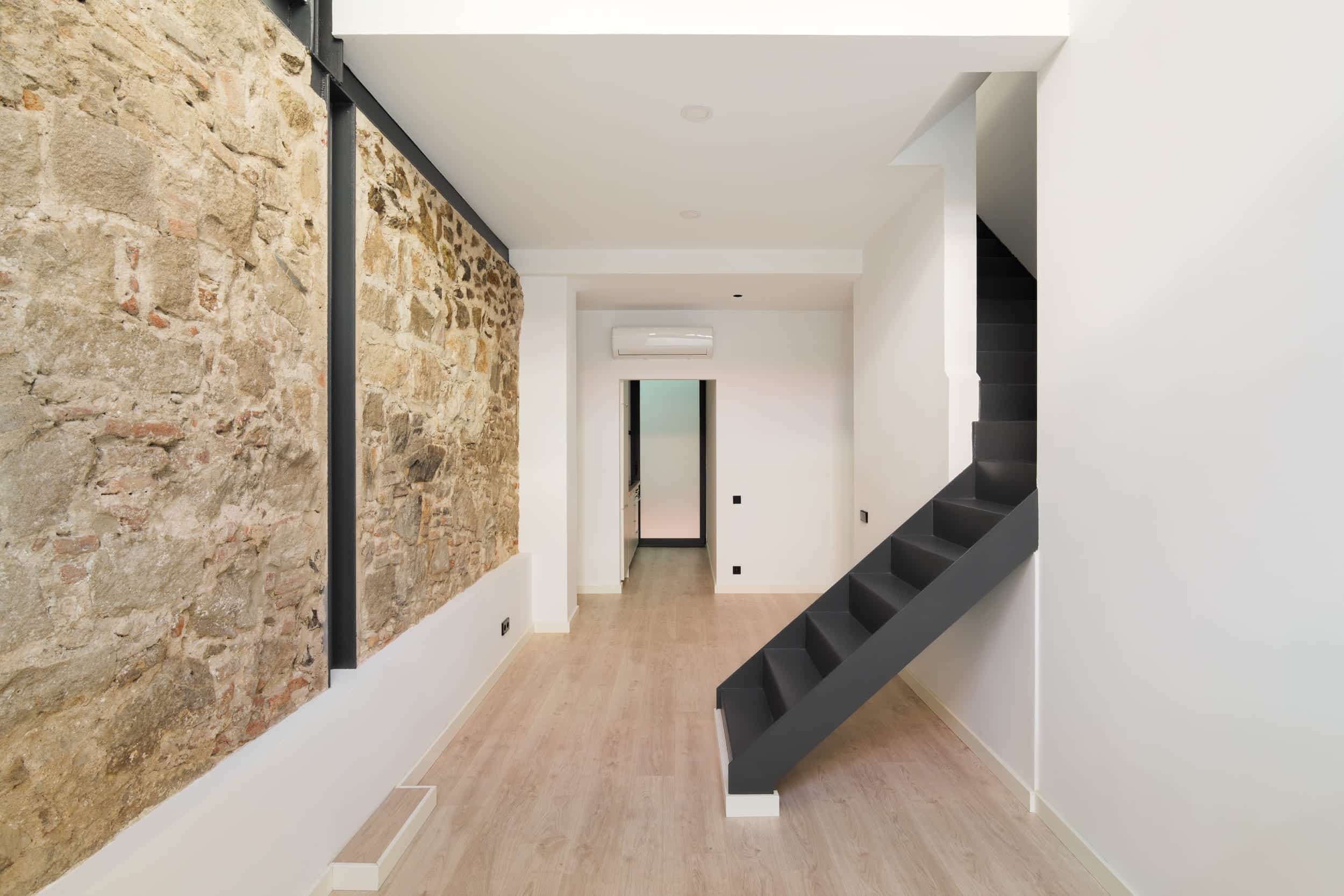 Reforma sostenible en una vivienda: hall y escalera, pared de piedra, suelo de madera y escalera en negro.